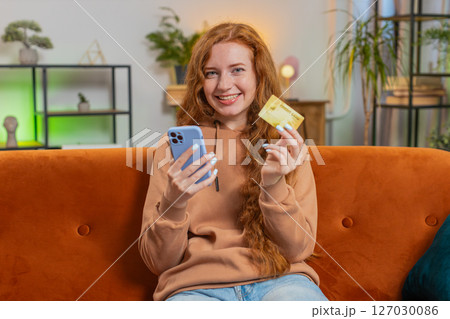 Happy young woman using credit bank card and smartphone while shopping online sitting on sofa couch Happy young woman using credit bank card and smartphone while shopping online sitting on sofa couch 127030086