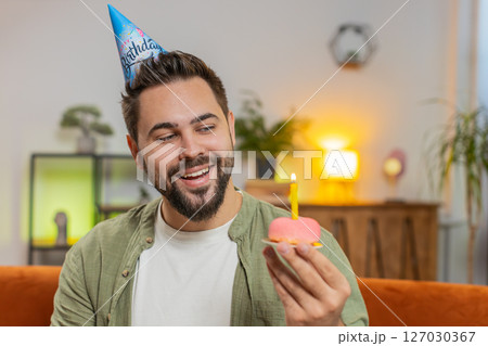 Happy young man wears birthday hat makes wish congratulating blowing burning candle on cake at home 127030367