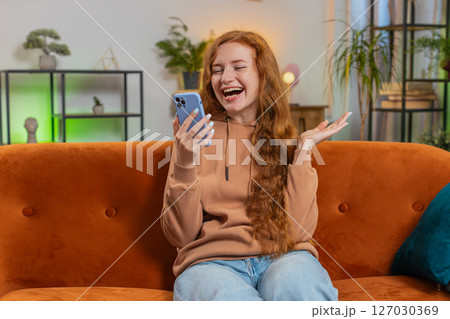 Happy young woman using cellphone celebrating success victory winning lottery on sofa in living room 127030369