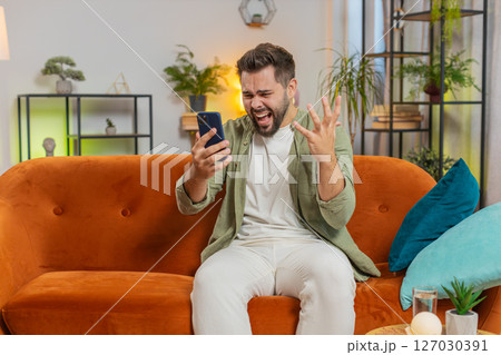 Upset displeased Caucasian man using smartphone reading bad news sitting on sofa in living room 127030391