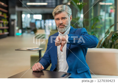 Upset Caucasian businessman in suit showing thumb down sign gesture sitting in modern office lobby 127030423