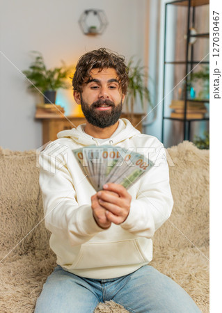 Successful rich young Hispanic man holding dollar bill money fan sitting on sofa couch at home 127030467
