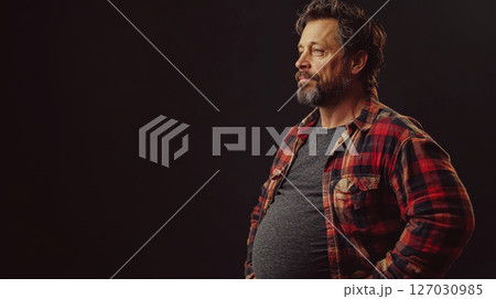 pregnant man in plaid shirt stands with hands on hips against dark background. Concept of confidence and casual style. 127030985