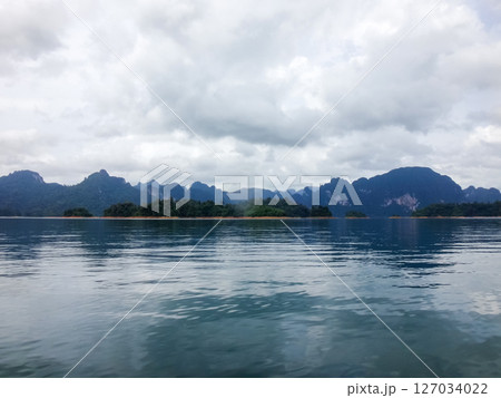 Tranquil Lake Vista: Capturing the serenity of a misty lake, surrounded by the majestic mountains and under a cloudy sky. The still waters reflect the atmospheric conditions. 127034022