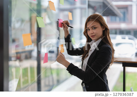 Businesswoman writing ideas on sticky notes on glass wall in office space. 127036387