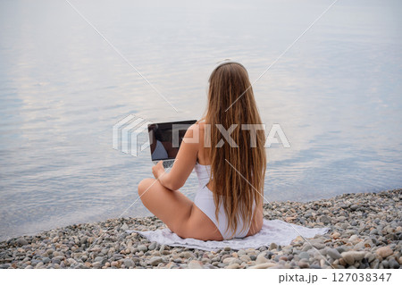 A woman is sitting on the beach with a laptop open in front of her. She is wearing a white bikini and has long hair. Concept of relaxation and leisure. 127038347