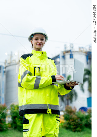 Vertical image of petrochemical or gas factory worker hold laptop for working and look at camera with smiling in front of the factory. 127044804