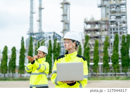 Petrochemical factory worker use walkie talkie to contact coworker with one coworker woman use laptop to work in front and they stay with factory in the background. Petrochemical factory worker use walkie talkie to contact coworker with one coworker woman use laptop to work in front and they stay with factory in the background. 127044817