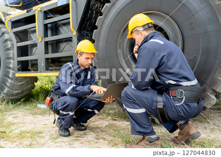 One factory worker use laptop and sit near wheel of truck and check or maintenance with his coworker in workplace area. One factory worker use laptop and sit near wheel of truck and check or maintenance with his coworker in workplace area. 127044830