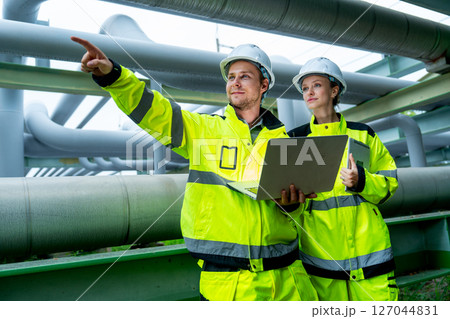 Two petrochemical or gas workers or technicians use laptop to work near pipe network with man also point to left direction. 127044831