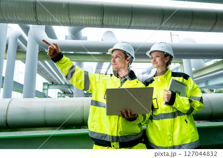 Side view of two petrochemical or gas workers or technicians use laptop to work near pipe network with man also point to left direction. Side view of two petrochemical or gas workers or technicians use laptop to work near pipe network with man also point to left direction. 127044832