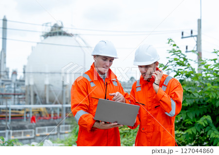 One engineer or gas factory worker point to laptop and discuss with the other one who use walkie-talkie and they stay in front of gas factory tank in workplace. 127044860