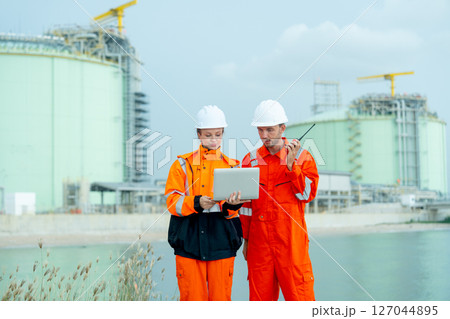 Two petrochemical or gas factory workers man and woman stand in front of factory tank near the bay and work together using laptop. Two petrochemical or gas factory workers man and woman stand in front of factory tank near the bay and work together using laptop. 127044895
