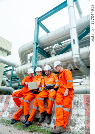 Vertical image group of petrochemical or gas factory workers or engineers discuss about work using laptop and stand near pipeline in factory workplace. Vertical image group of petrochemical or gas factory workers or engineers discuss about work using laptop and stand near pipeline in factory workplace. 127044918