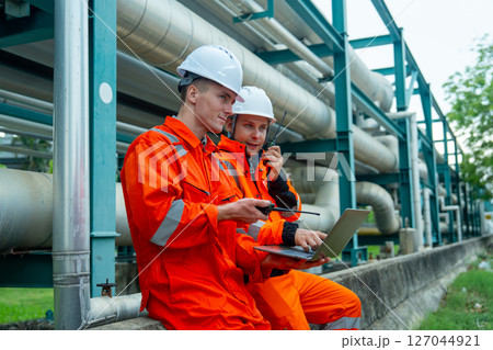 Two petrochemical or gas factory workers or technicians man adiscuss about work using laptop and sit near pipeline in factory workplace. 127044921