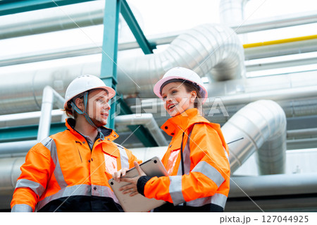 Two petrochemical or gas factory workers man and woman discuss about work using laptop and stand near pipeline in factory workplace. 127044925