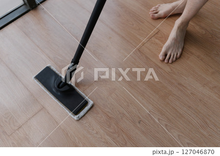 Top view of unrecognizable barefoot man washing hardwood floor with steam cleaner mop in modern apartment, ensuring hygiene and cleanliness. Concept of modern housekeeping appliance. 127046870