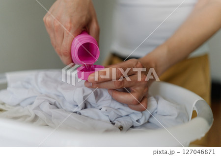 Male hands pouring stain remover from pink bottle onto white clothes soaking in plastic basin, preparing laundry for washing or soaking process, close-up . Concept of household chores, daily routines. 127046871