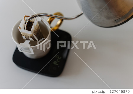 Barista preparing drip coffee using kettle, precision kitchen scales and drip coffee bag, carefully pouring hot water over coffee bag placed in cup on scales, close-up, creating morning ritual. Barista preparing drip coffee using kettle, precision kitchen scales and drip coffee bag, carefully pouring hot water over coffee bag placed in cup on scales, close-up, creating morning ritual. 127046879