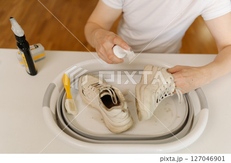High-angle view of cleaning white sneakers with scrubbing brush and detergent, removing stubborn stains from footwear in basin, preserving shoe hygiene and appearance thorough washing work. 127046901