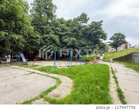 Empty playground with swings and seesaws in a quiet residential area. Peaceful atmosphere of childhood spaces surrounded by summer greenery. 127047756