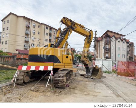 Excavator parked on construction street. Urban infrastructure, civil engineering, and machinery maintenance in residential neighborhoods Excavator parked on construction street. Urban infrastructure, civil engineering, and machinery maintenance in residential neighborhoods 127049114