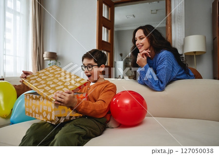 Excited child opens gift with mother nearby in cozy living room setting Excited child opens gift with mother nearby in cozy living room setting 127050038