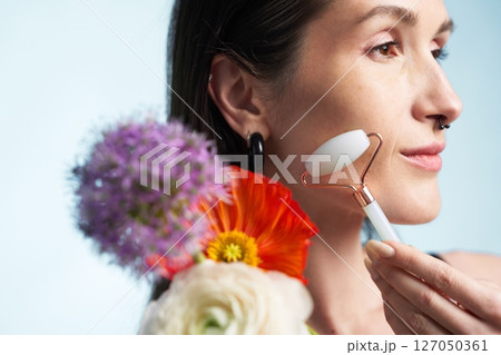 Woman using jade roller for skincare with flowers at a bright location 127050361