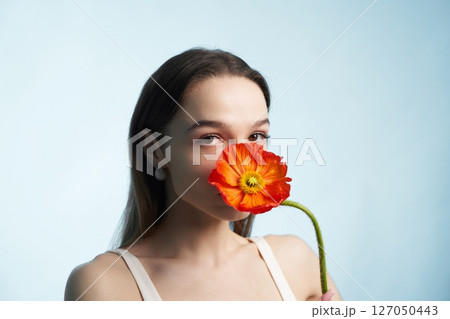 Young woman holds an orange flower near her face against a blue background Young woman holds an orange flower near her face against a blue background 127050443