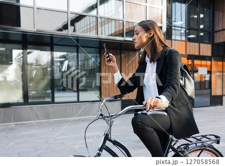 Businesswoman sitting on bicycle and checking her mobile phone. Young female in formal clothes using mobile phone while sitting on bicycle. 127058568
