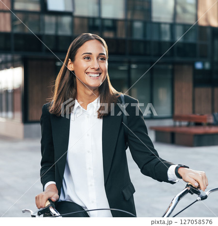 Happy female entrepreneur standing outdoors with bicycle at business building. Portrait of a smiling businesswoman wearing formal wear and a backpack holding bicycle. Happy female entrepreneur standing outdoors with bicycle at business building. Portrait of a smiling businesswoman wearing formal wear and a backpack holding bicycle. 127058576