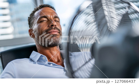 Man enjoying cool breeze from fan in office during extremely hot weather conditions 127066437