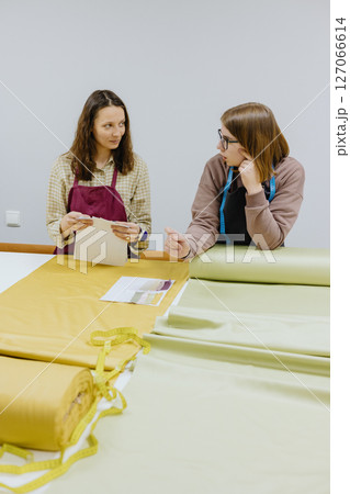 Two female seamstresses and fashion designers stand at table with roll of fabric negotiate Two female seamstresses and fashion designers stand at table with roll of fabric negotiate 127066614