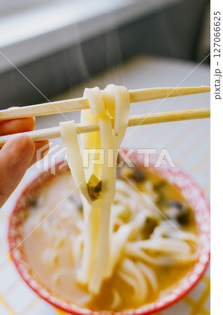 person is eating noodles with chopsticks. colorful bright red plate of ramen sits on yellow table. person is eating noodles with chopsticks. colorful bright red plate of ramen sits on yellow table. 127066625