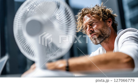 Man enjoying cool breeze from fan in bright room during extremely hot weather conditions 127066779