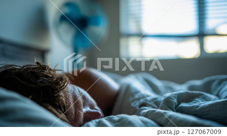 Sweaty individual sleeping in bed with fan nearby, struggling to cope with extreme heat 127067009