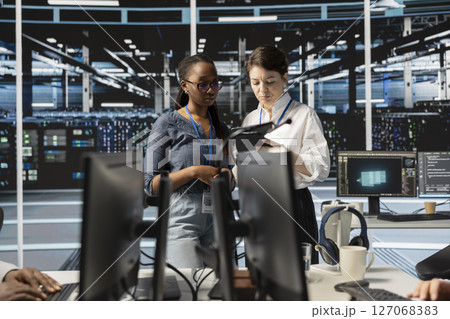 Team of technicians using tablet in server farm, analyzing data. Women working in data center examining infrastructure using device, ensuring system integrity and security 127068383