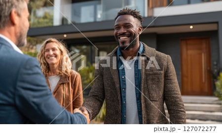 Happy real estate agent shaking hands with new homeowners front of modern house, celebrating successful property deal with smiles and positive 127072277