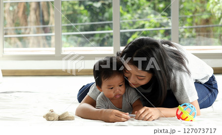 Mother lovingly embraces her baby from behind as they lie on soft white blanket while toddler holds teether and focuses calmly near large window view 127073717
