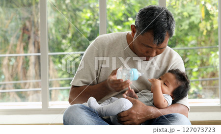 Father cradling baby on lap while holding feeding bottle, care and attention in quiet indoor moment near window 127073723