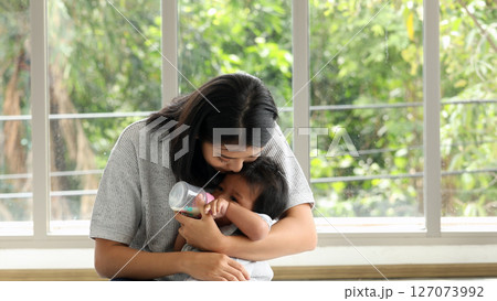 Mother gently kissing baby head while bottle-feeding in front of bright windows during bonding time 127073992