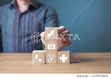 A person arranging wooden blocks with healthcare icons including a syringe, stethoscope, cross, and medication, symbolizing medical care, health services, and wellness solutions on a blue background. A person arranging wooden blocks with healthcare icons including a syringe, stethoscope, cross, and medication, symbolizing medical care, health services, and wellness solutions on a blue background. 127074845