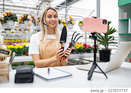 A Caucasian woman, working in a florist shop, presents gardening tools while streaming online from a table with a laptop and smartphone. A Caucasian woman, working in a florist shop, presents gardening tools while streaming online from a table with a laptop and smartphone. 127077737