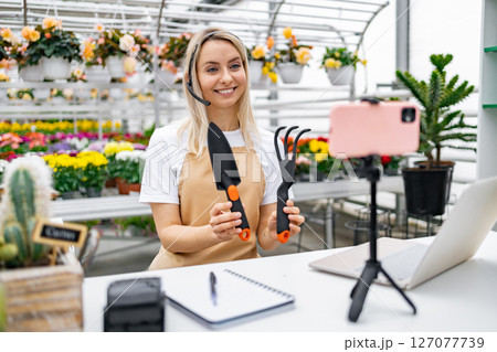 A Caucasian woman, a florist, explains gardening tools in her shop, preparing for a live sale, with a friendly and inviting demeanor. A Caucasian woman, a florist, explains gardening tools in her shop, preparing for a live sale, with a friendly and inviting demeanor. 127077739