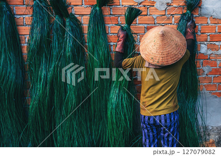 Vietnamese woman in traditional straw hat dries dyed green reeds at a woven mat factory in a village in Vietnam 127079082