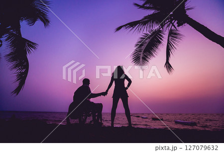 Disabled man in a wheelchair with his wife on the beach. Silhouettes at sunset Disabled man in a wheelchair with his wife on the beach. Silhouettes at sunset 127079632