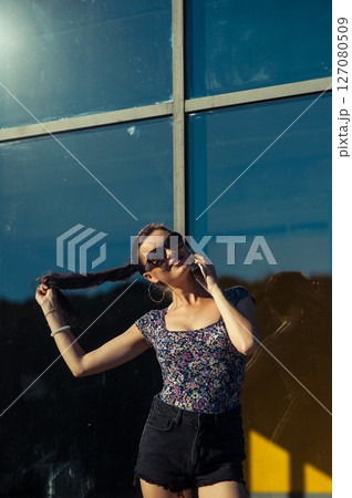 A young woman with glasses and a cell phone in her hands is having a conversation near a glass structure 127080509