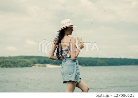 A young girl stands by the lake with a bright cup of juice in her hand A young girl stands by the lake with a bright cup of juice in her hand 127080523