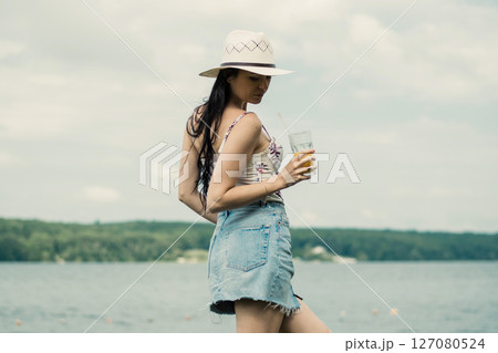 A young girl stands by the lake with a bright cup of juice in her hand A young girl stands by the lake with a bright cup of juice in her hand 127080524