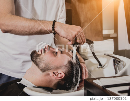 Young man with trendy haircut at barber shop. Barber washes customer head. Young man with trendy haircut at barber shop. Barber washes customer head. 127080591